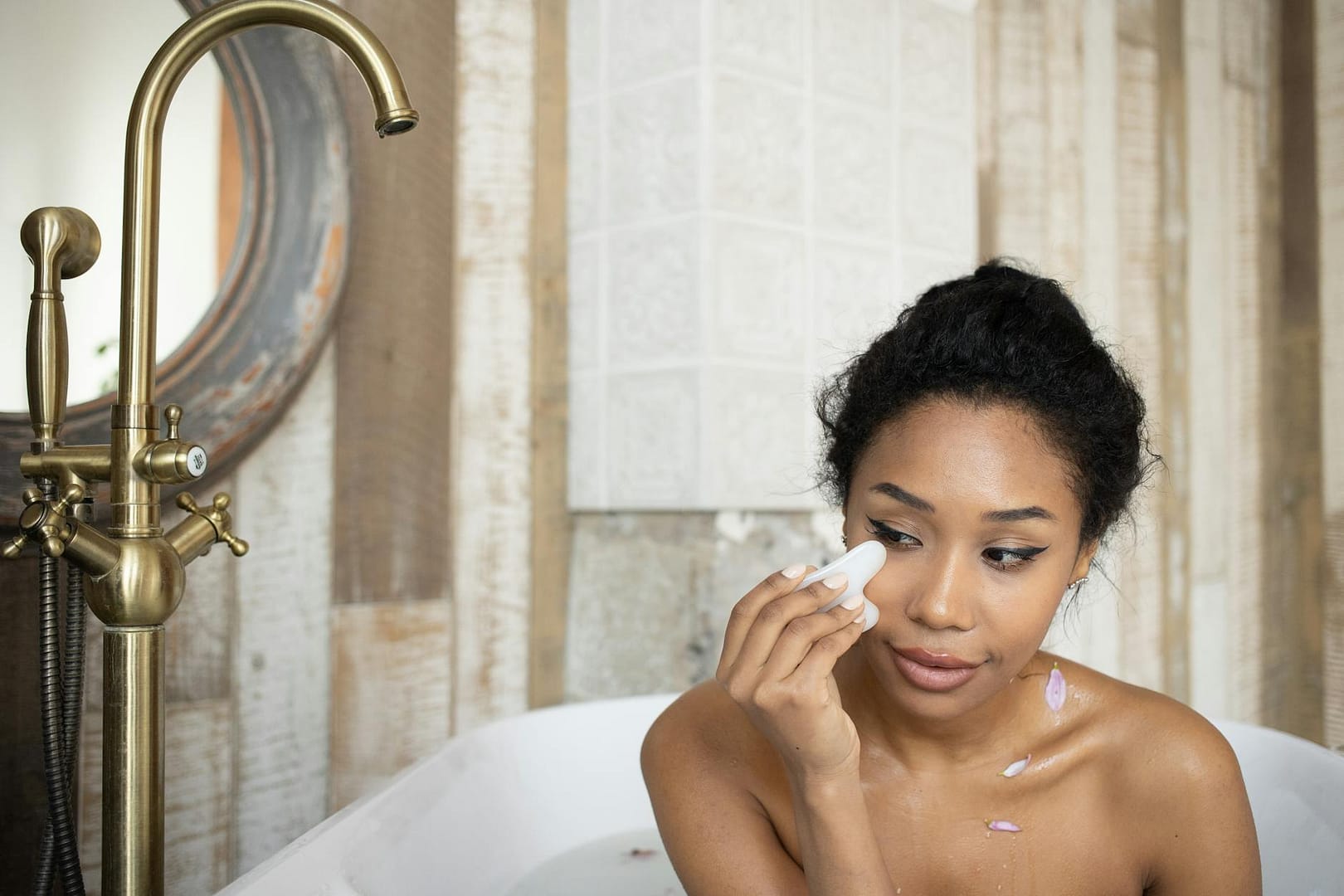 black woman doing facial massage in bathtub with metal faucet